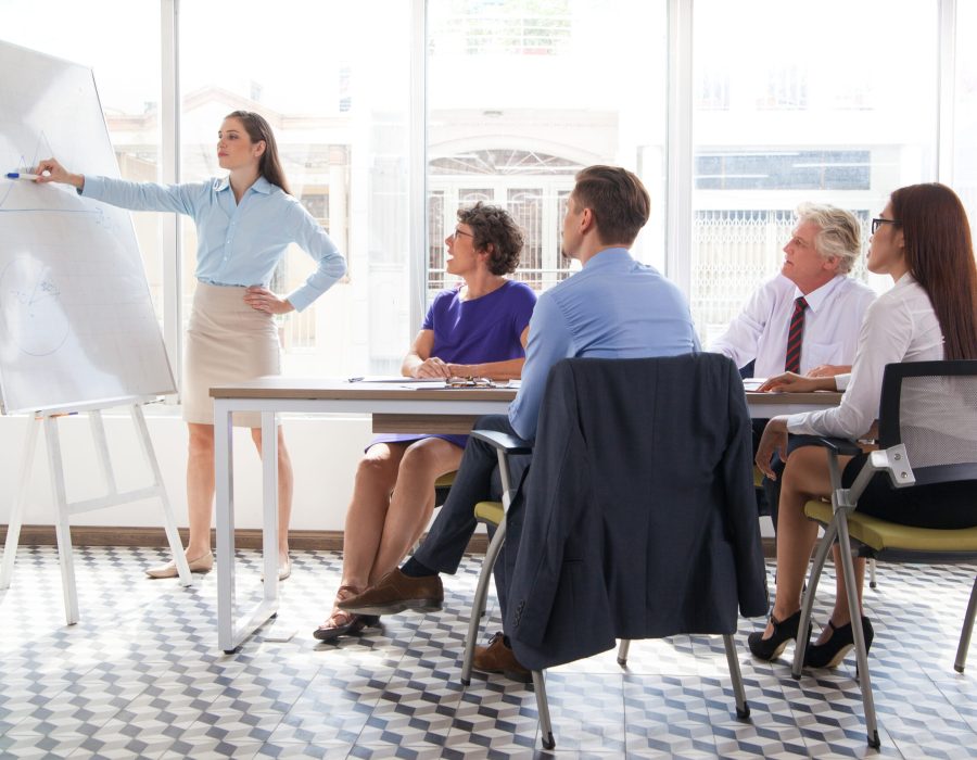 Confident female business coach pointing at whiteboard and explaining graph at meeting, group of four people listening to her. They sitting in conference room. Business training or education concept